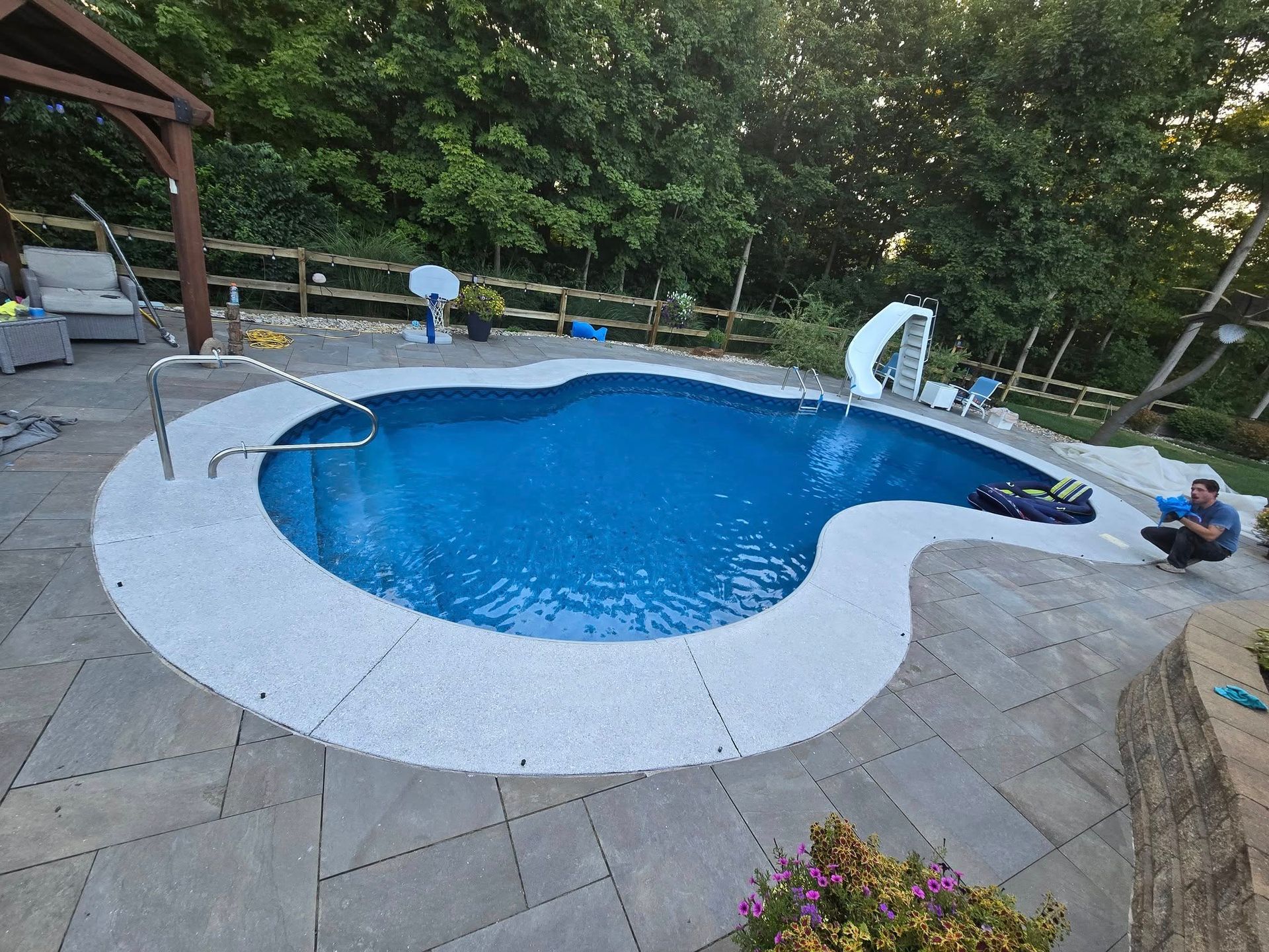 A kidney-shaped swimming pool with blue water, surrounded by a white border and stone patio, with a slide and a person near it.