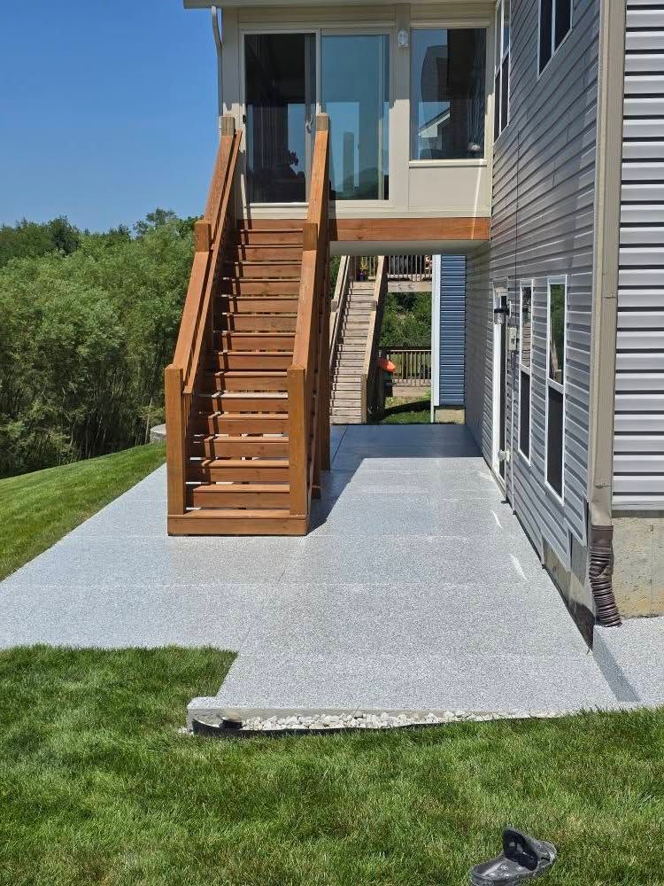 Patio with concrete surface next to a house with wooden stairs leading up. Green grass surrounds.