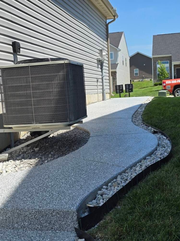 Pathway of speckled concrete curving along a lawn, next to a building and an AC unit.