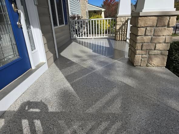 Front porch with gray aggregate concrete. Blue door on the left, white railing, and stone columns on right.