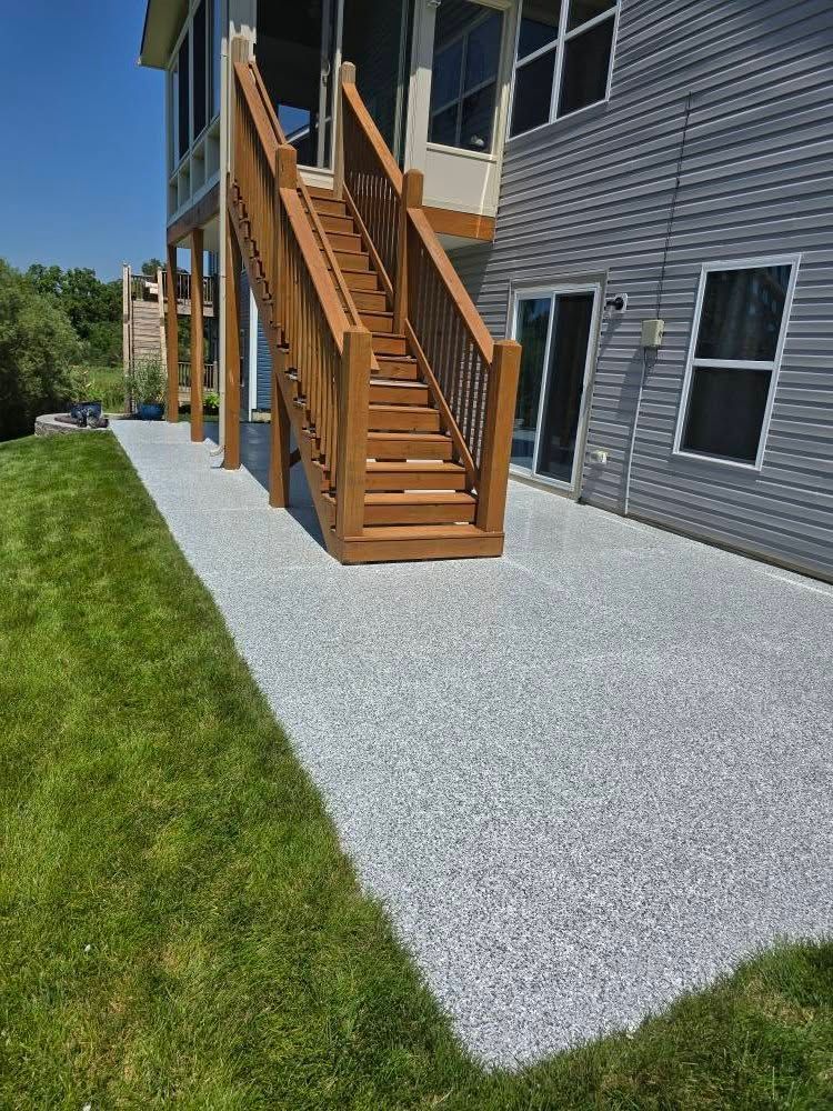 Gray pebble-like patio next to a wooden staircase and a light gray house, with green grass in view.