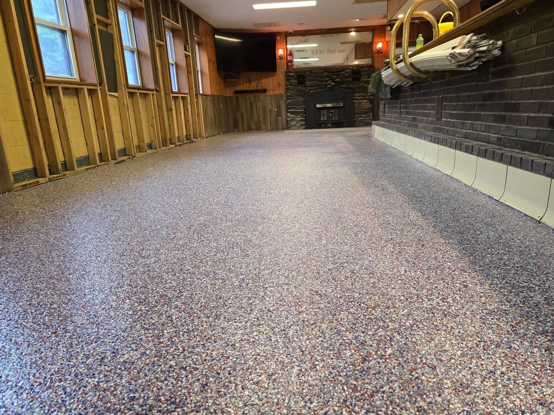 Interior view of a room with a speckled epoxy floor, bar, exposed wall studs, and a stone fireplace.