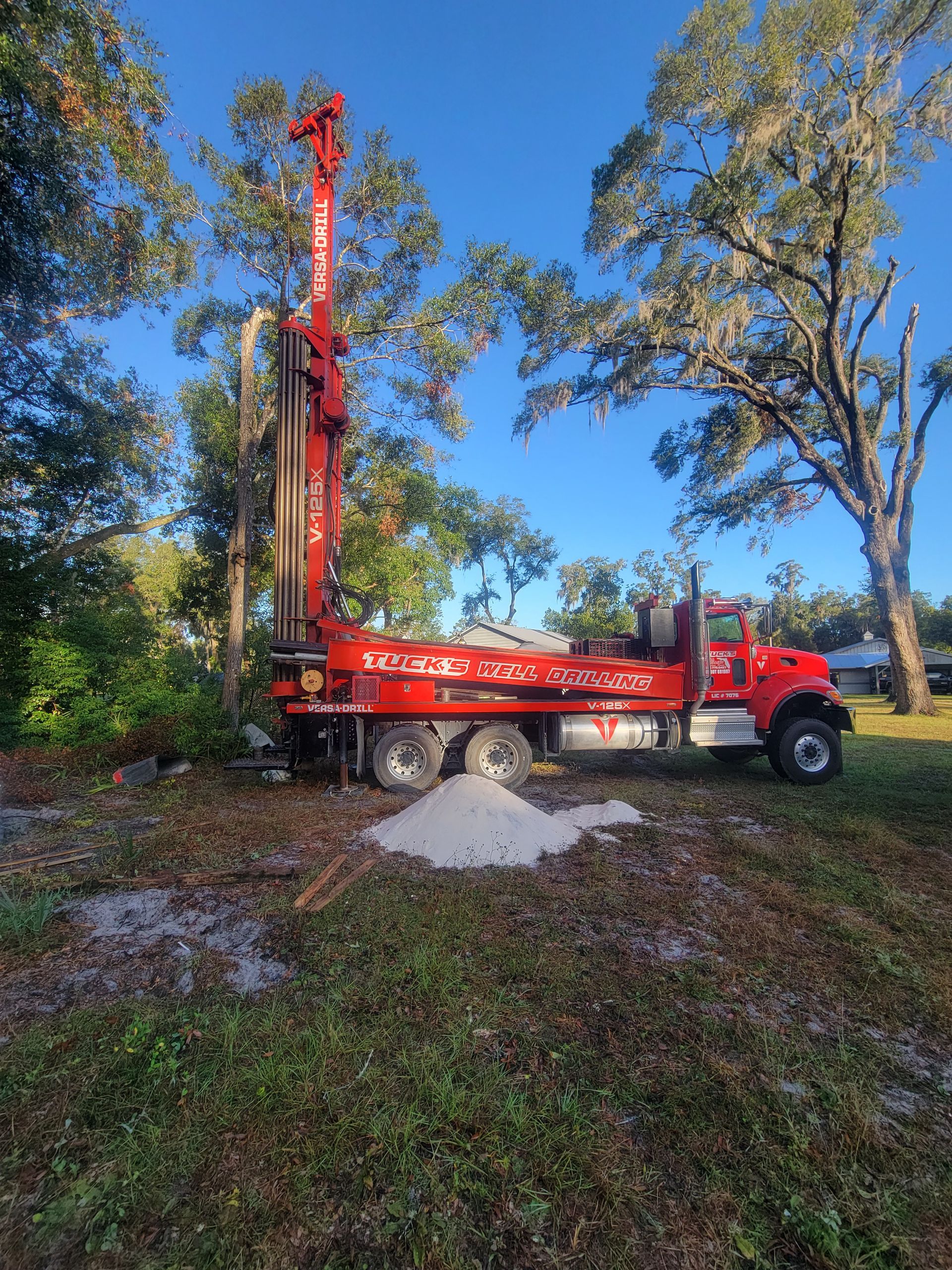 Red drilling truck with tall auger on a grassy dirt site among trees under a blue sky