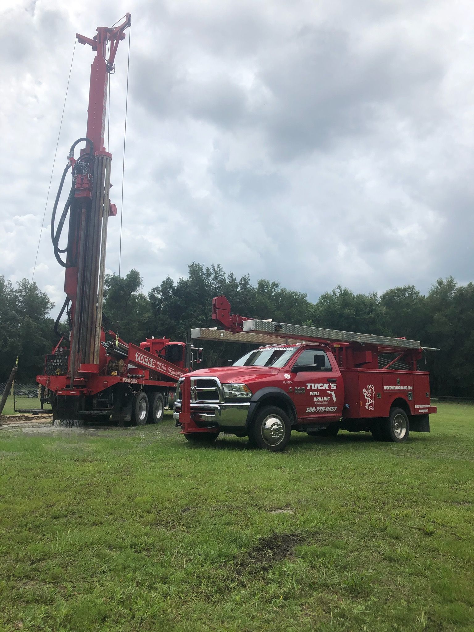 Red drilling rig and red utility truck on a grassy field under cloudy sky