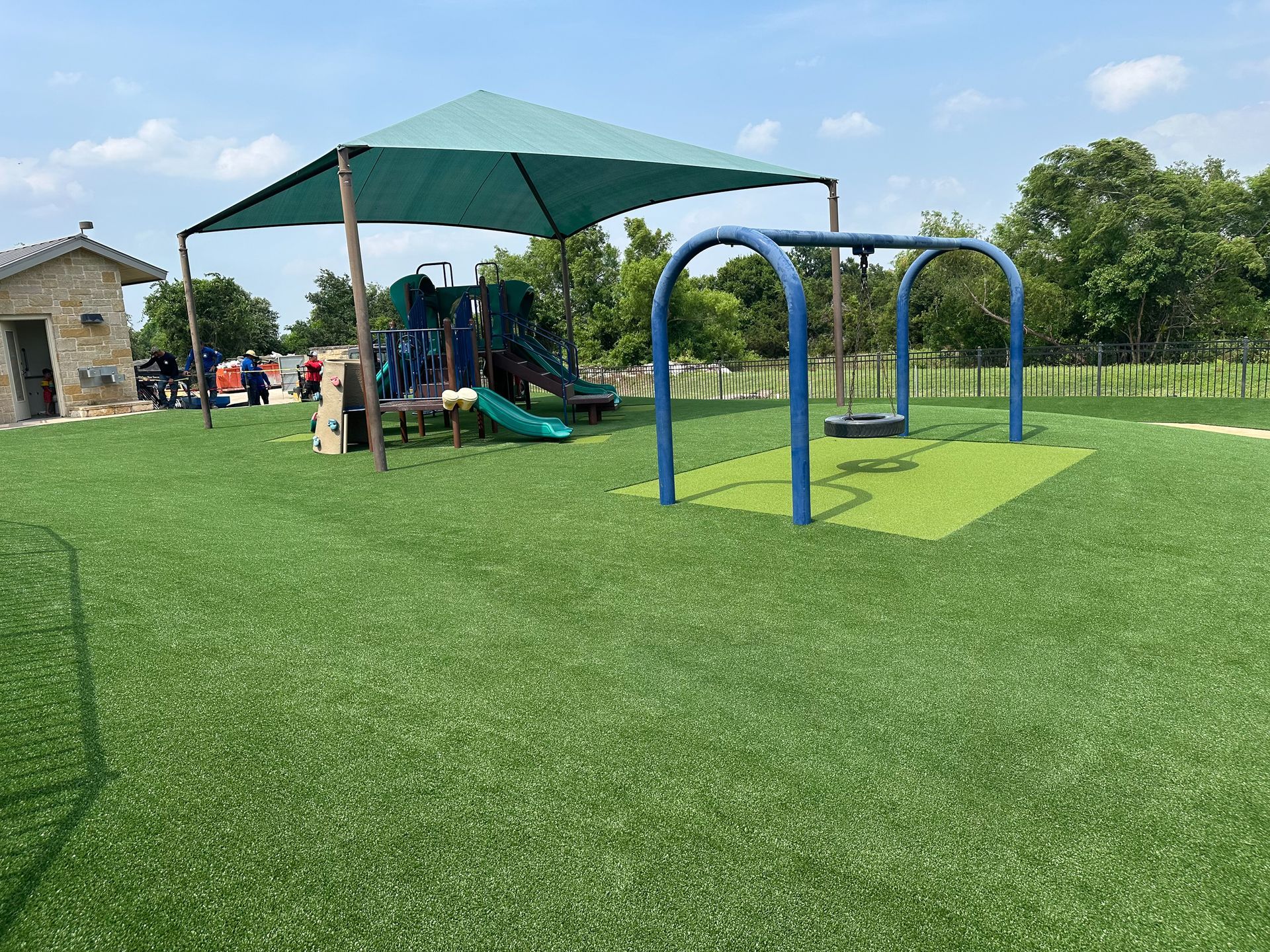 Playground with green turf, shaded play structure, swing set, and a small building.