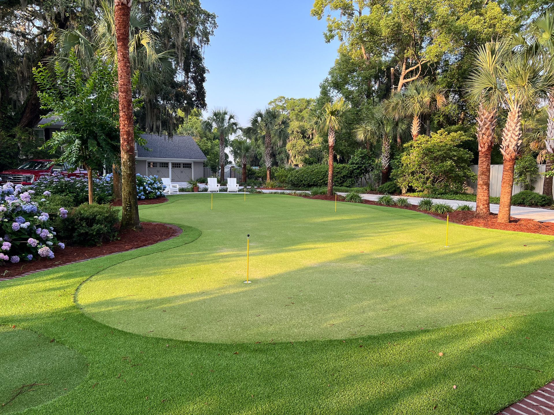 Lush green putting green with three holes, surrounded by landscaping, trees, and a small building in the background.