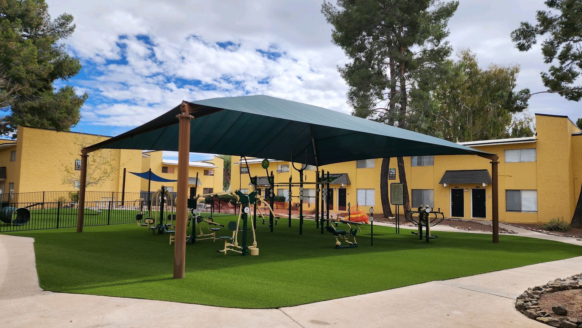 Outdoor fitness area under a large shade structure. Exercise equipment on green turf. Yellow building in background.