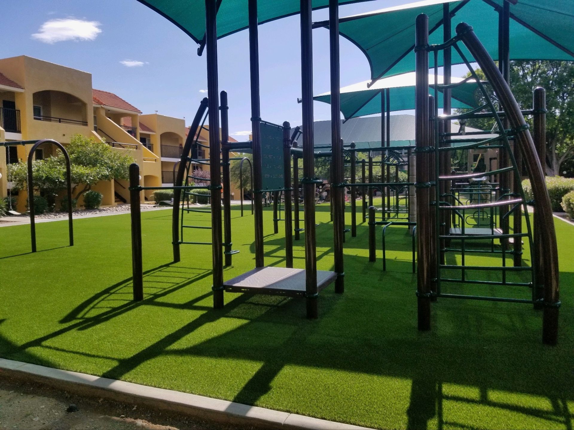 Playground with climbing structures, shaded by turquoise canopies, on green turf. Buildings in background.