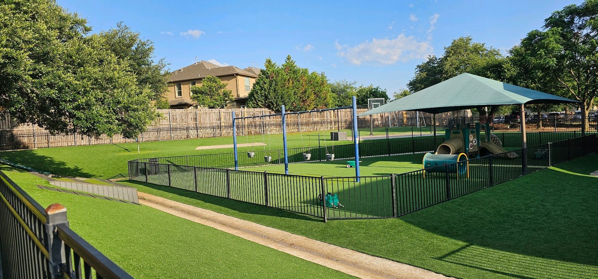 A dog park with green turf, play equipment, and a covered seating area. Trees and a building are in the background.