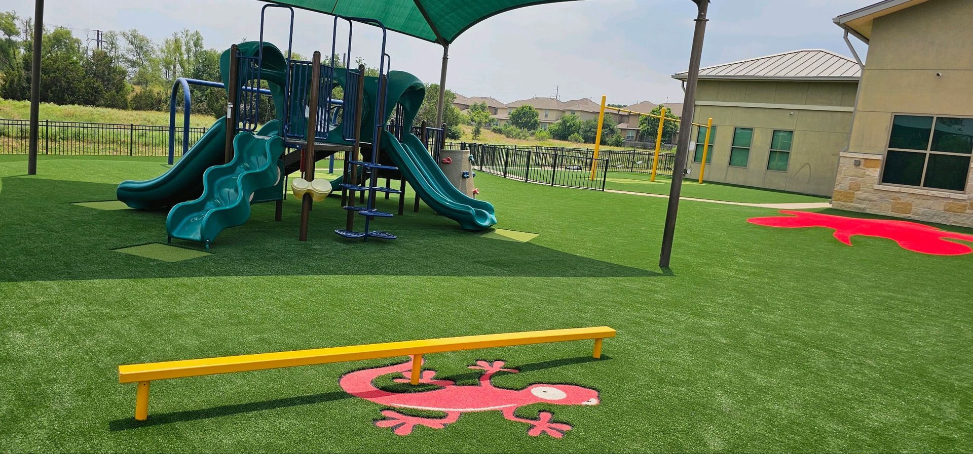 Playground with slides, balance beam, and green artificial turf. Building with windows in background.