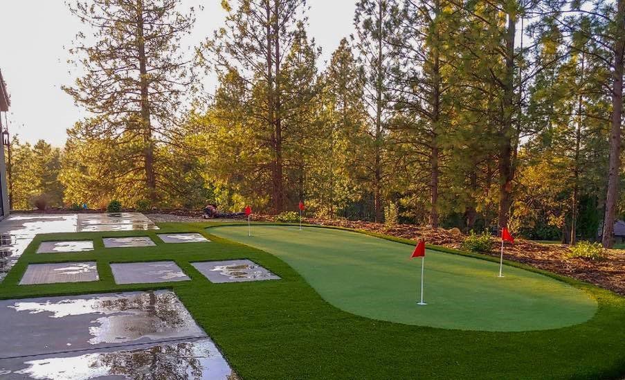 Green putting green with red flags, next to a stone patio, surrounded by trees.
