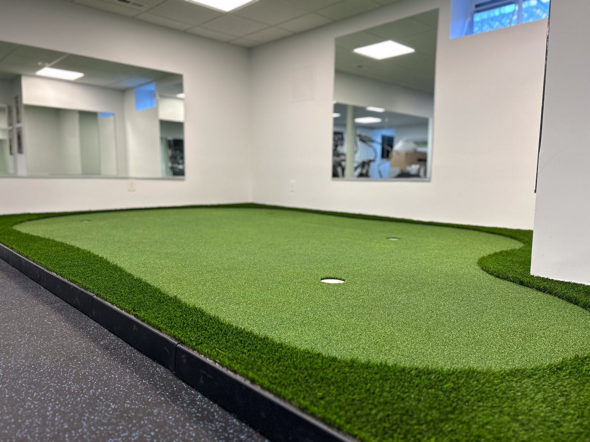 Indoor putting green with white walls, mirrors, and a dark border, next to a rubber floor.
