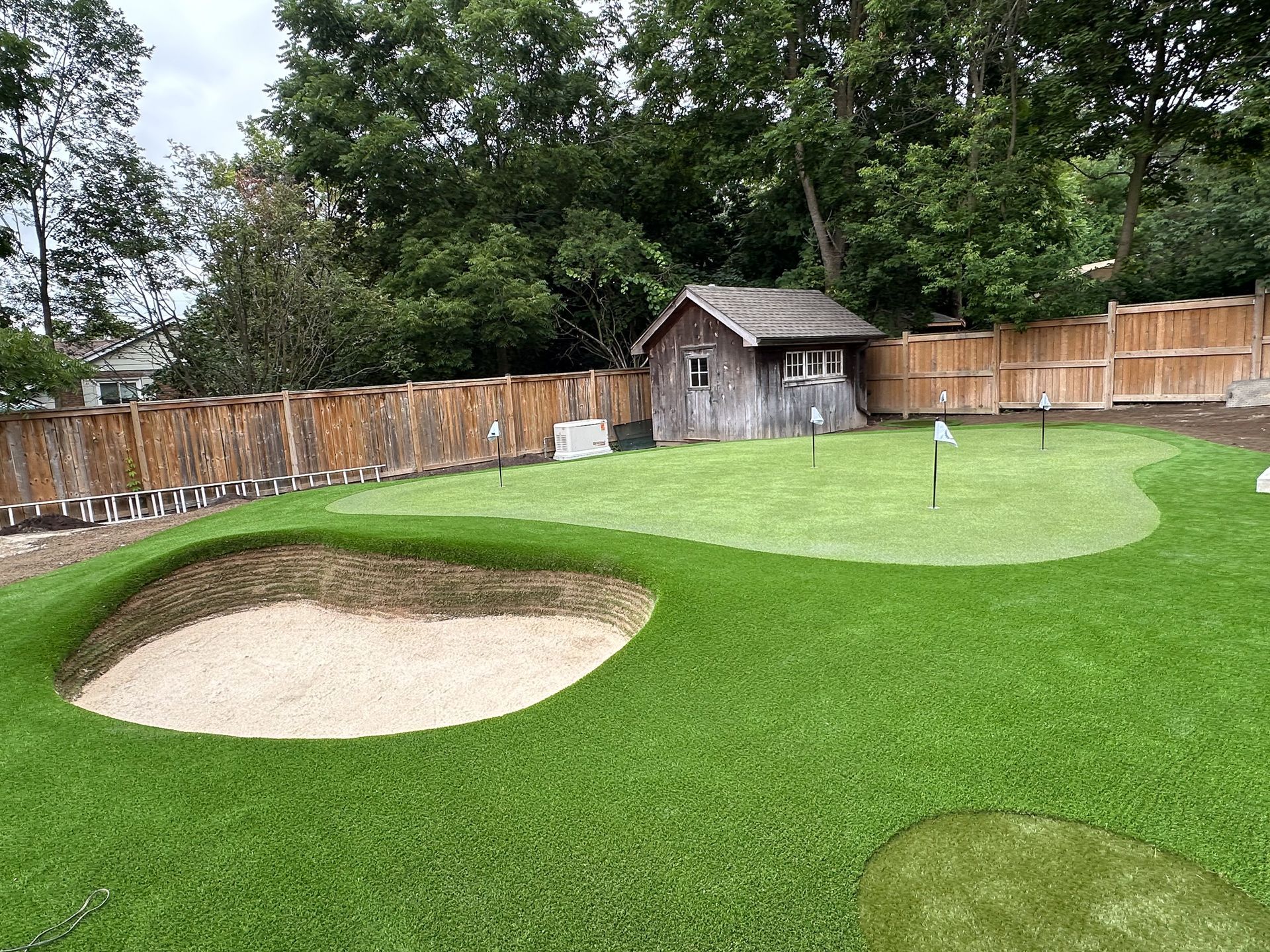 A backyard putting green with a sand trap, small wooden shed, and wooden fence.