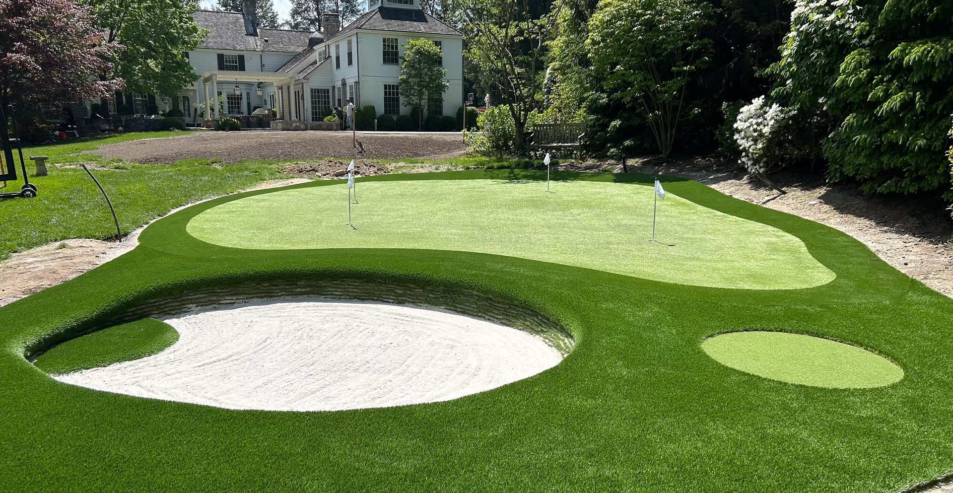 Backyard putting green with sand trap, flagsticks, and house in the background. Green grass and white sand.