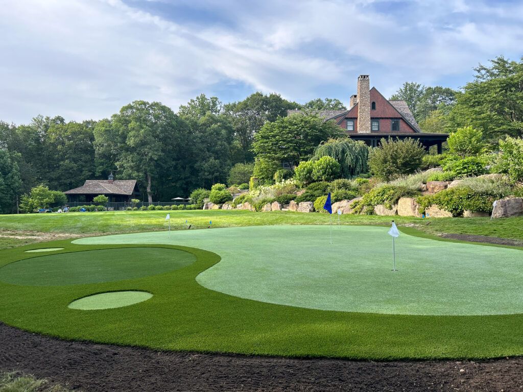 A putting green in front of a large house, surrounded by landscaping and trees.