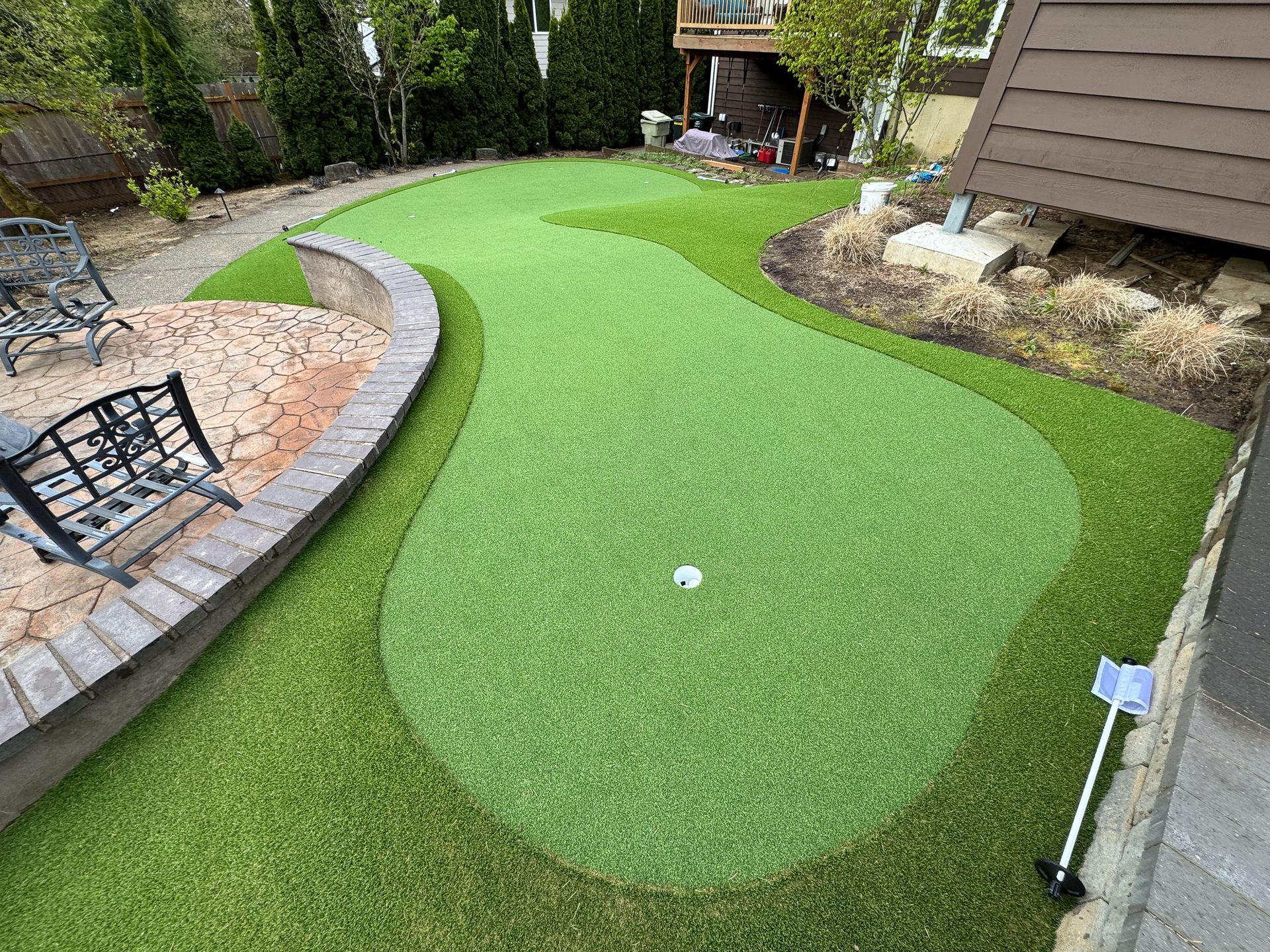Backyard putting green, bright green turf, white golf ball, brick border, patio furniture.