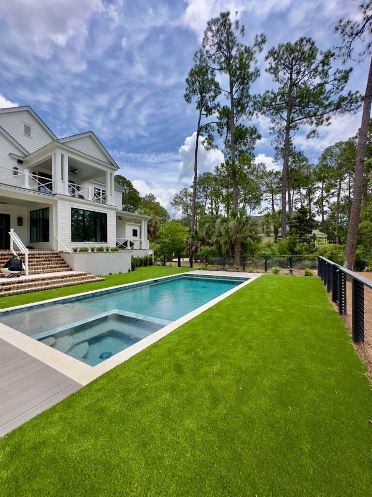White house with pool; green lawn; tall trees, blue sky.