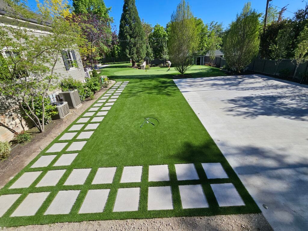 Backyard with stone pavers, green turf, and concrete driveway. Trees and building in background.