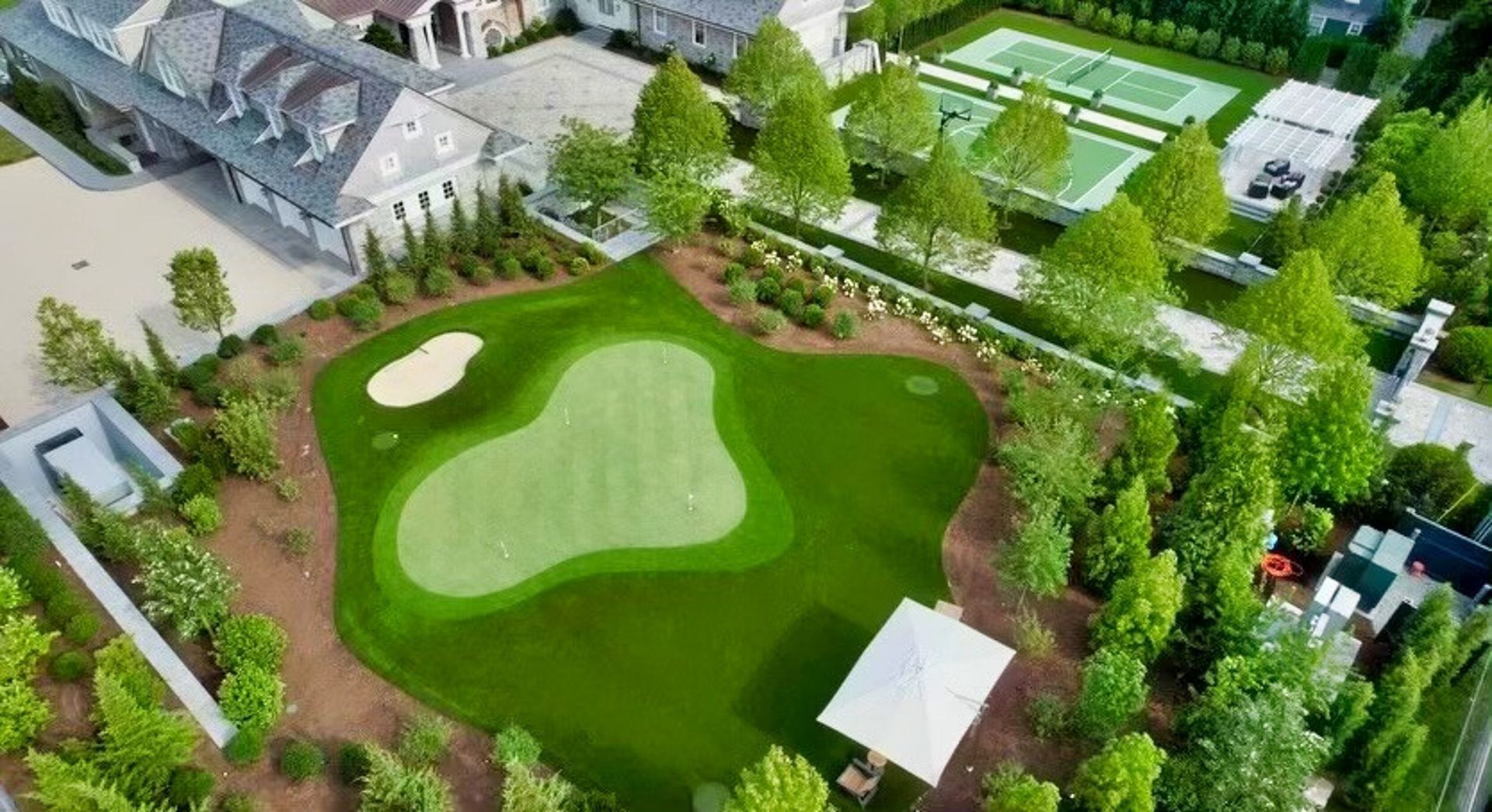 Aerial view of a private golf green, sand trap, and small white gazebo, surrounded by landscaping and buildings.