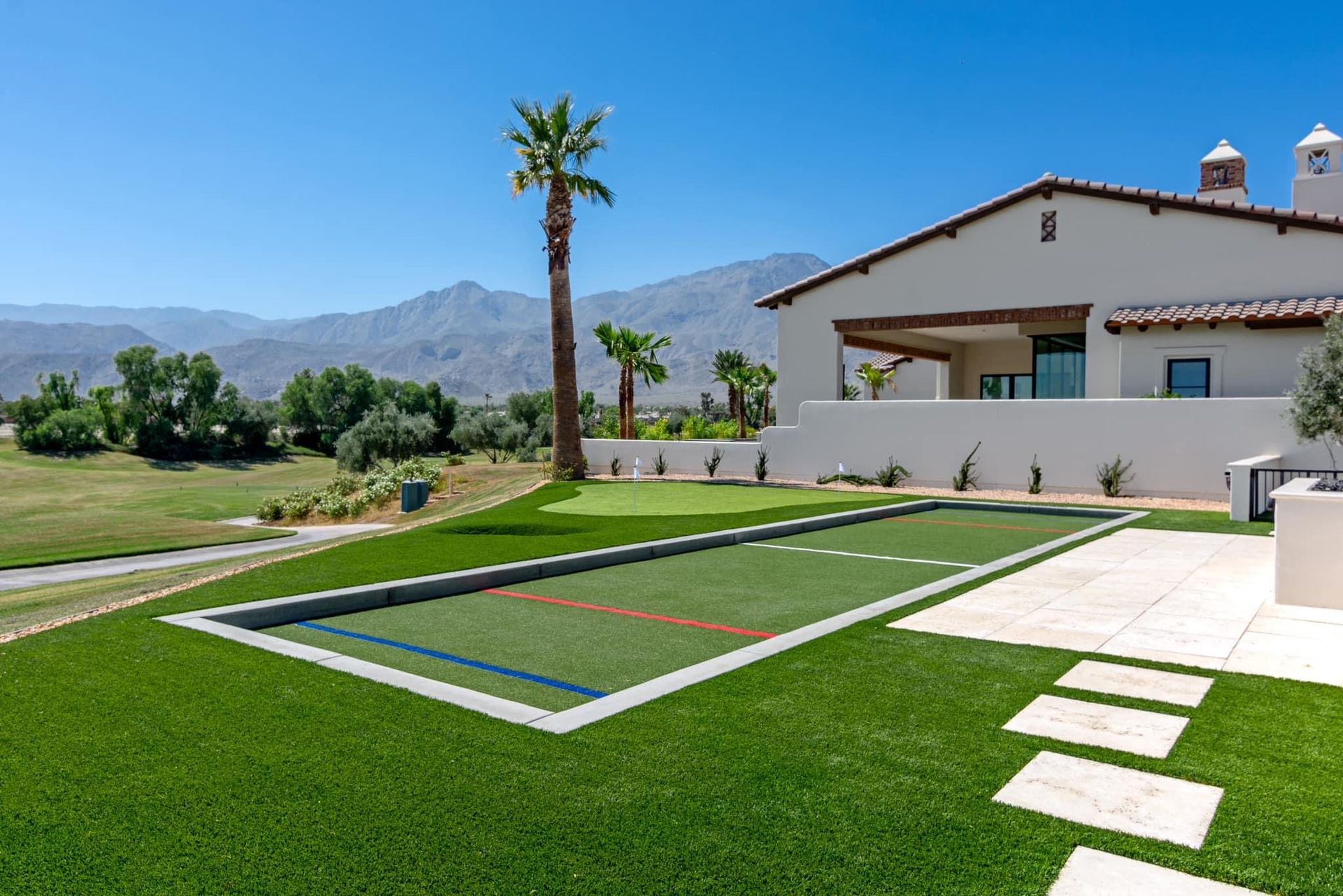 Bocce ball court on green artificial turf next to a house with mountain backdrop and palm tree.