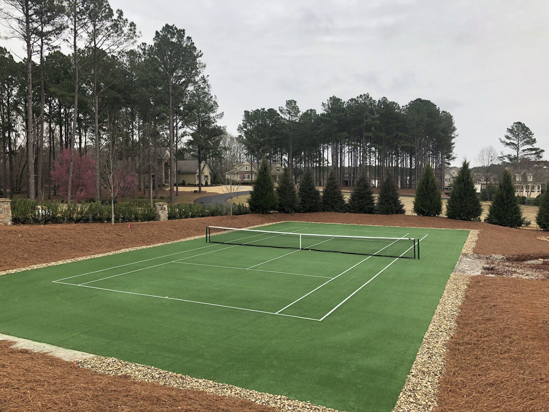 Tennis court with green artificial turf, surrounded by pine straw and evergreens, under an overcast sky.