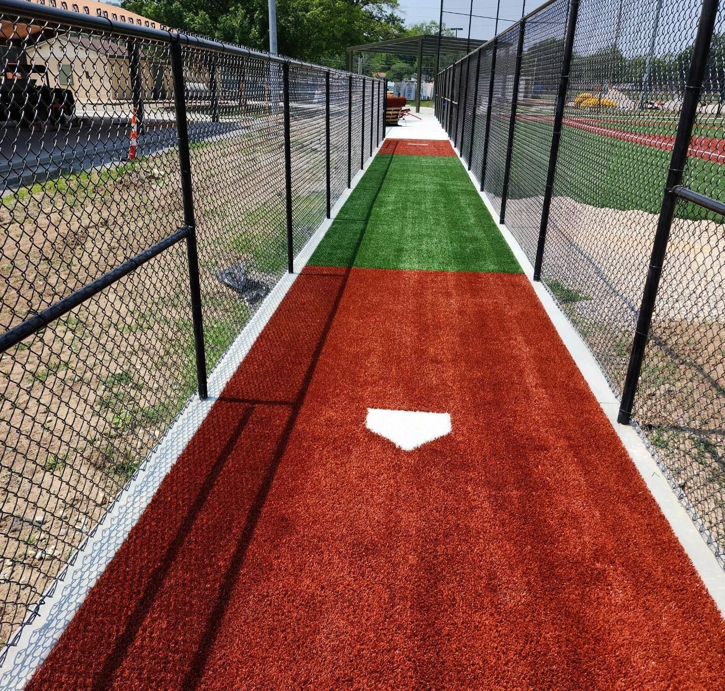 A turf baseball dugout pathway with red and green sections, white home plate, and chain-link fences.