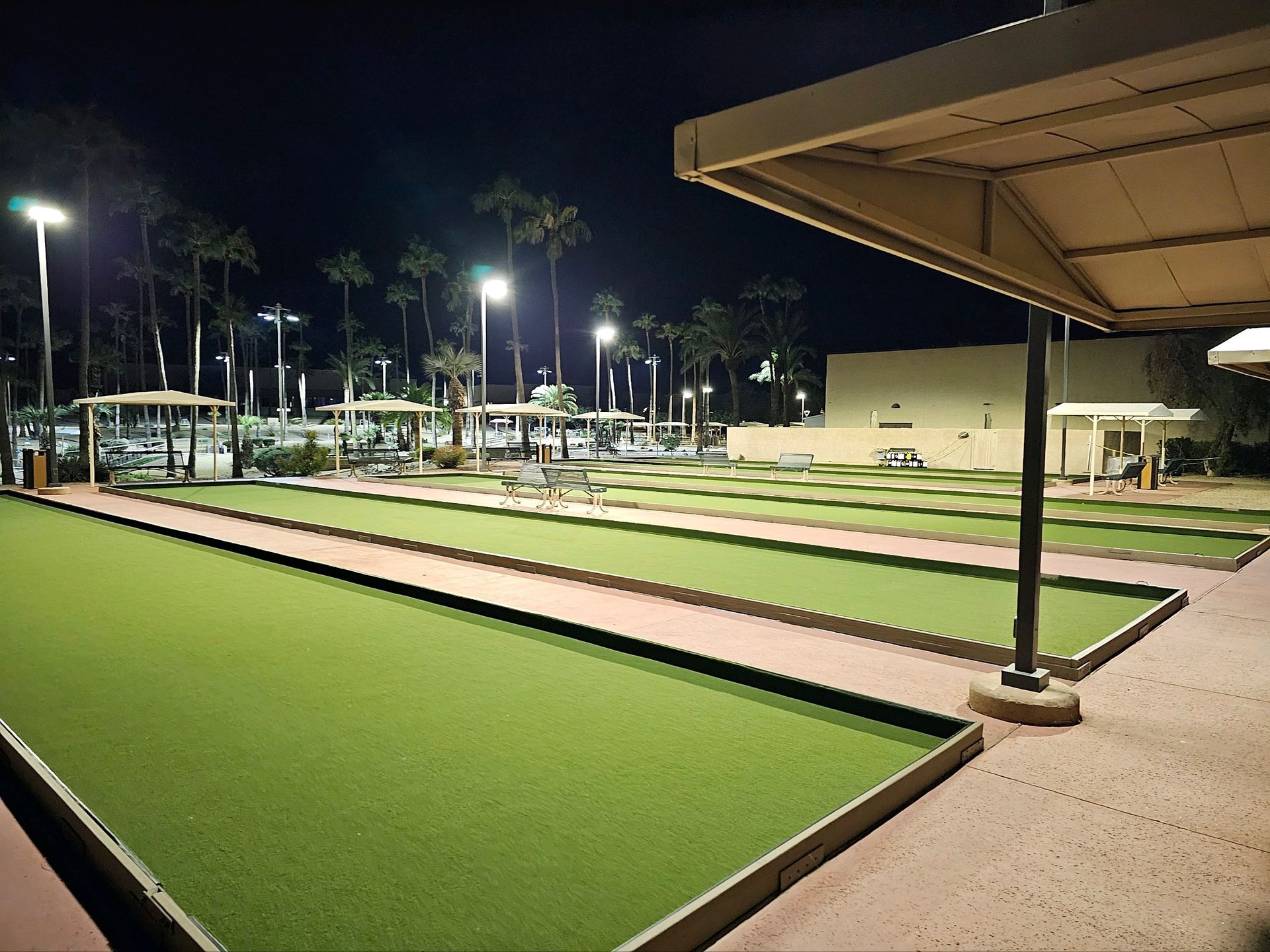 Night view of illuminated bocce ball courts with covered seating and palm trees.