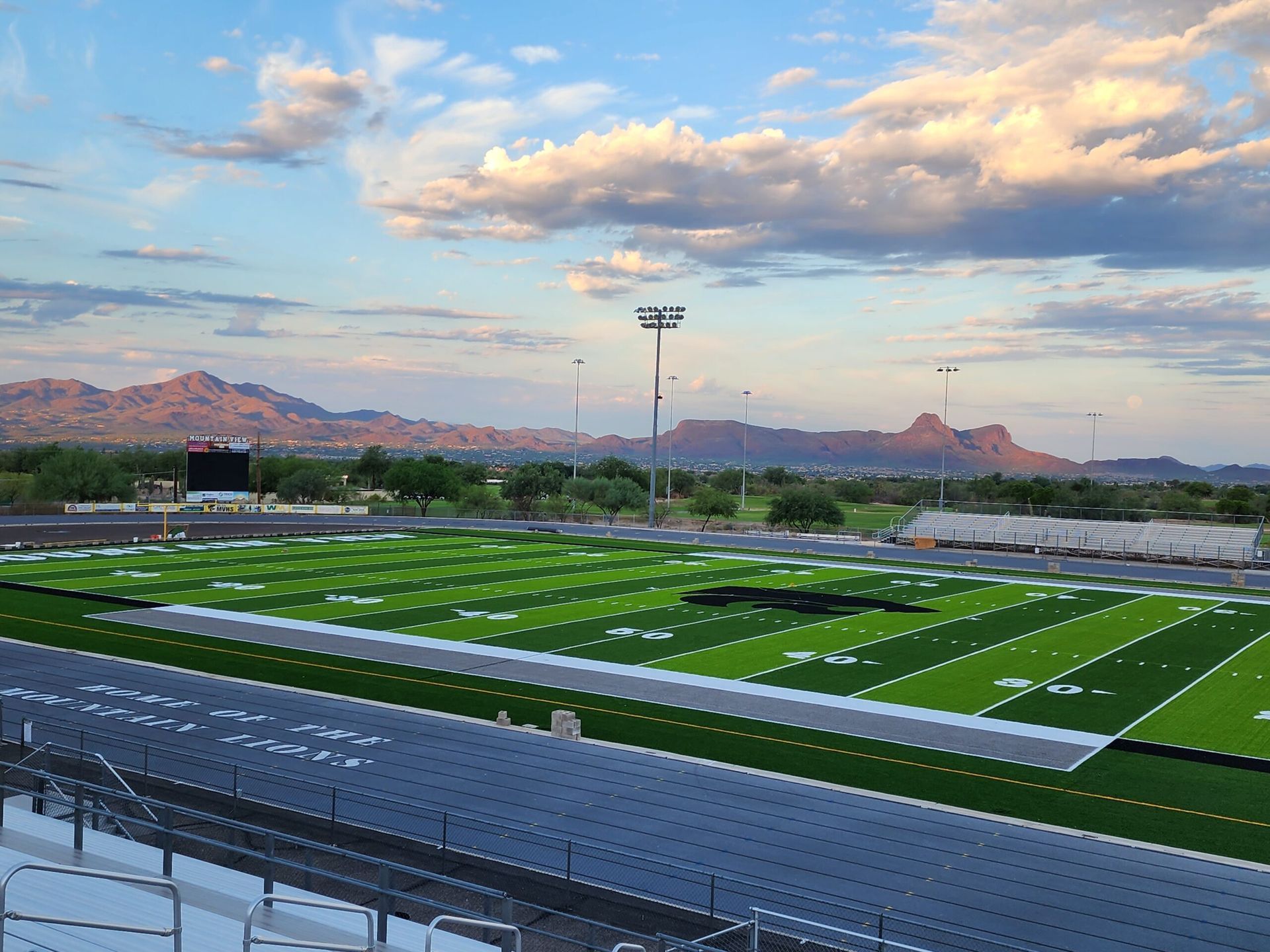 Football field under a cloudy sky with mountains in the background. Green grass and bleachers.