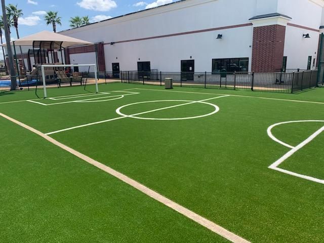A small soccer field with white lines on green turf, next to a building and fenced area.