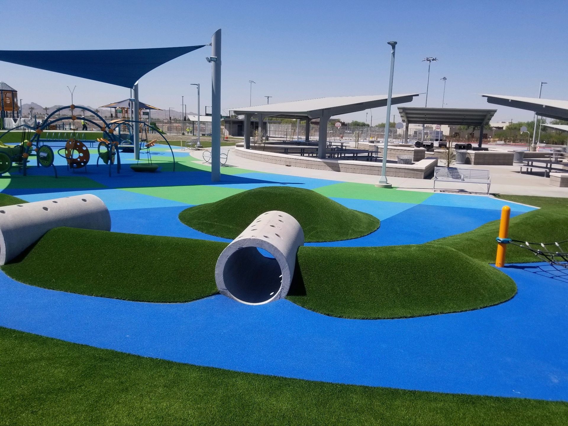 Playground with blue, green, and artificial turf, tunnels, hills, and a shaded structure.