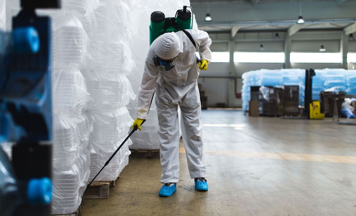 A worker in a white hazmat suit and protective gear sprays disinfectant on pallets of goods in a large warehouse.