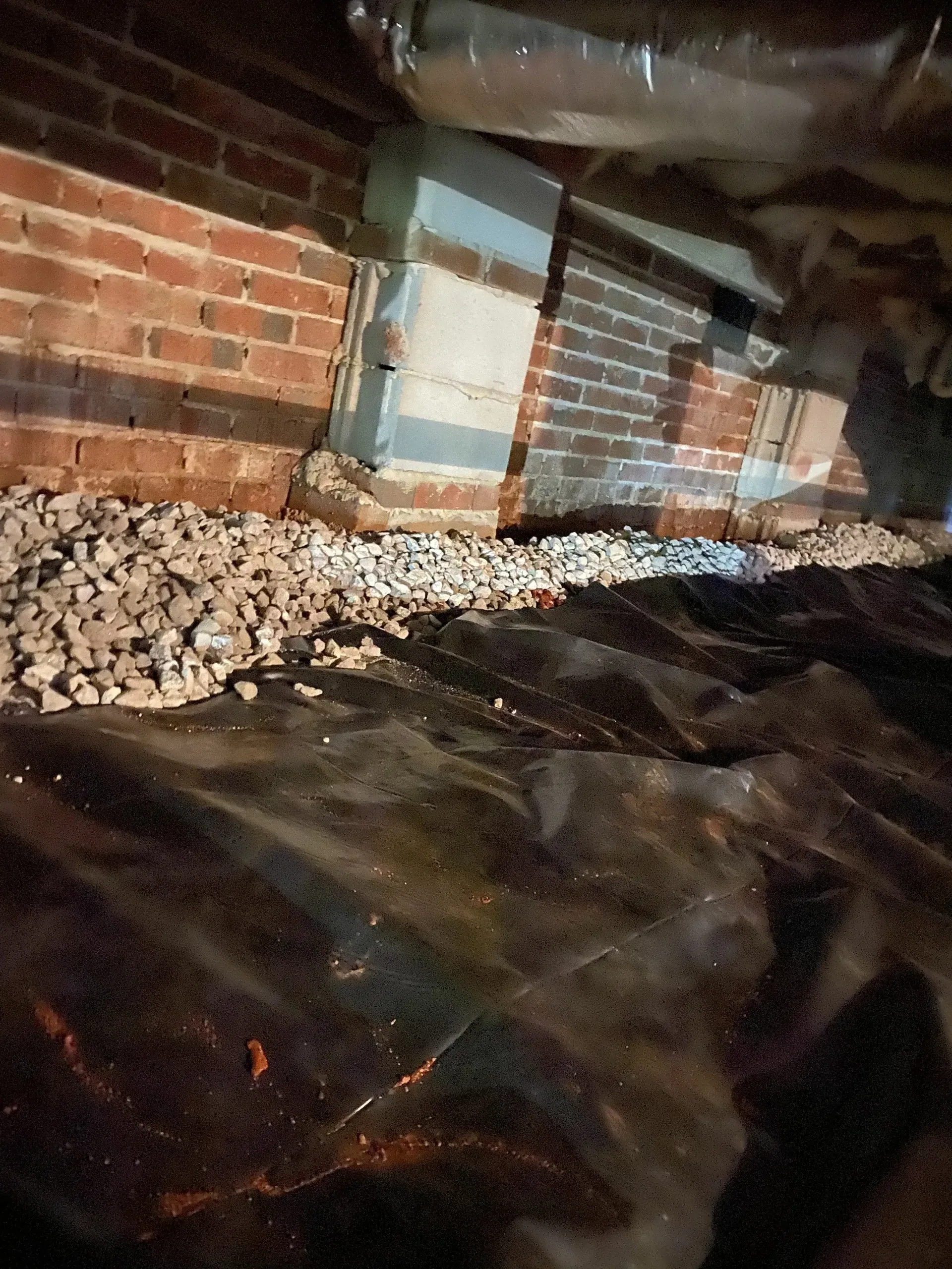 A crawl space with brick walls, gravel, and black plastic sheeting on the ground.