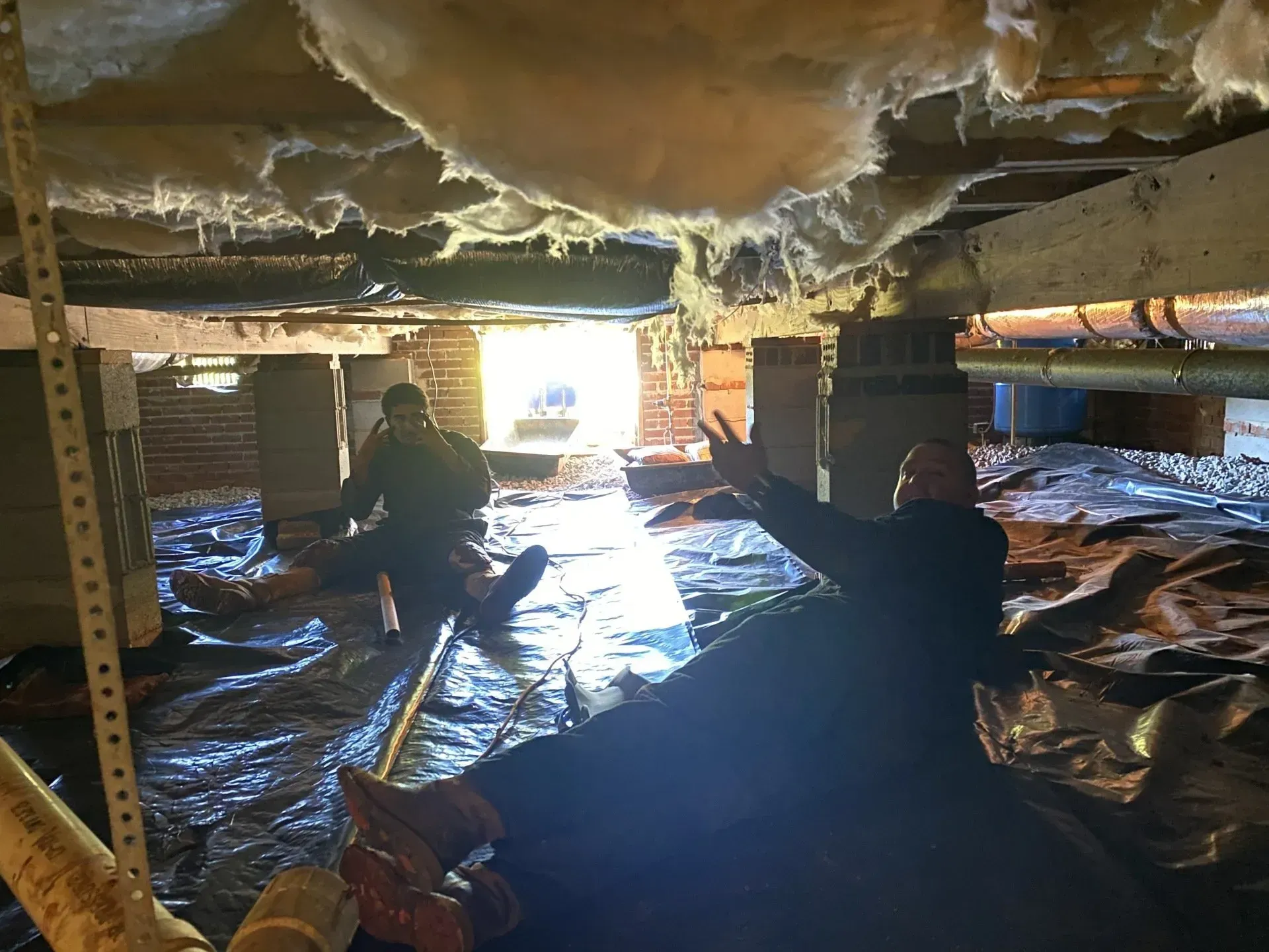 Two people working in a crawl space under a house, possibly doing repairs; black plastic on floor, insulation overhead.