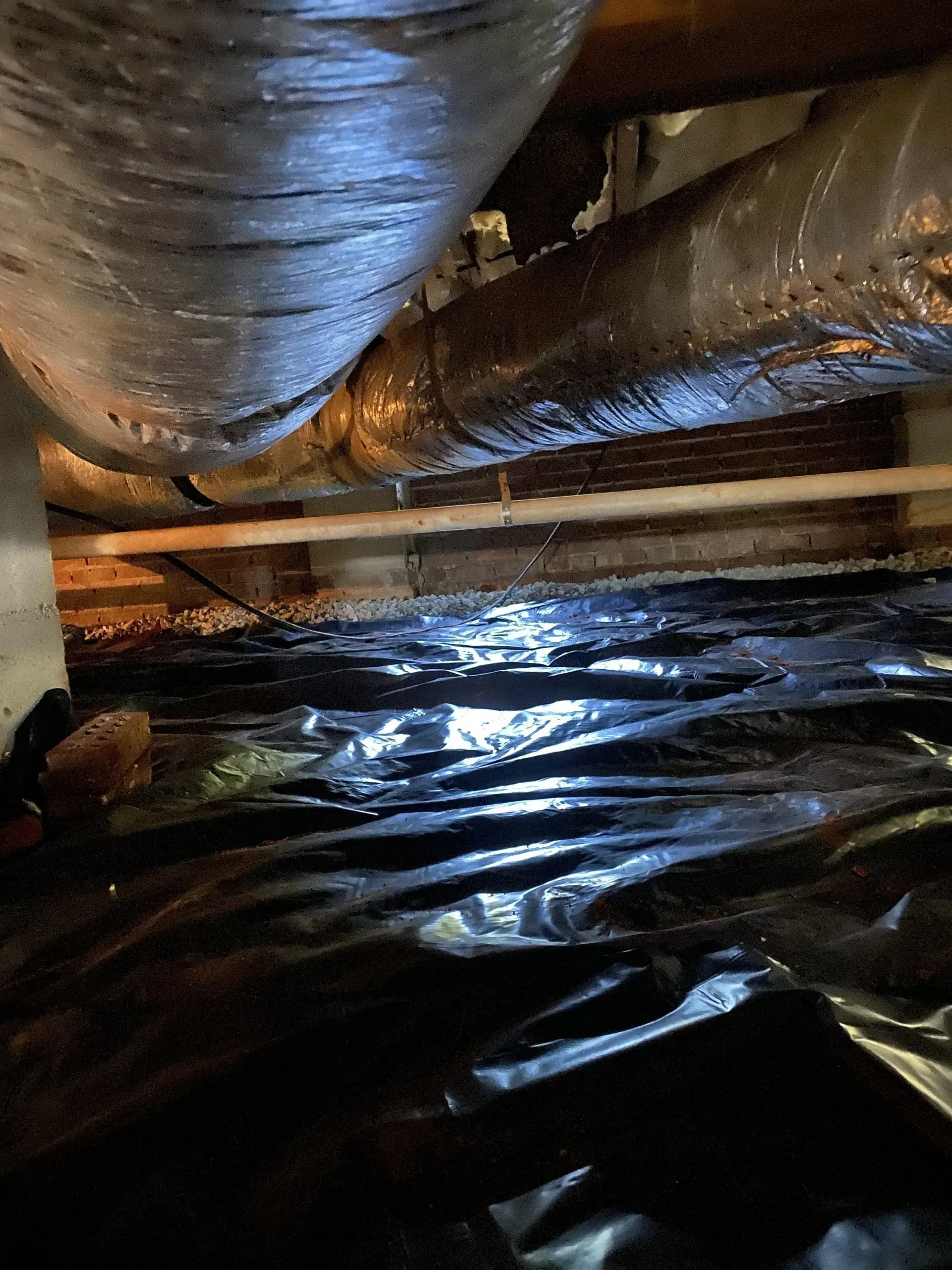 Under-house view showing HVAC ducting, wood beams, and black plastic sheeting covering the ground.