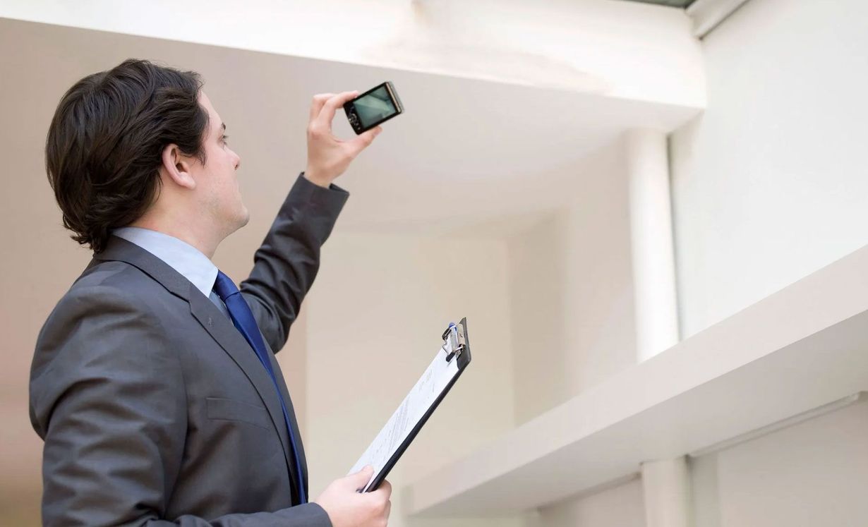 A professional in a suit holds a clipboard and uses a smartphone to take a photo of an indoor ceiling.