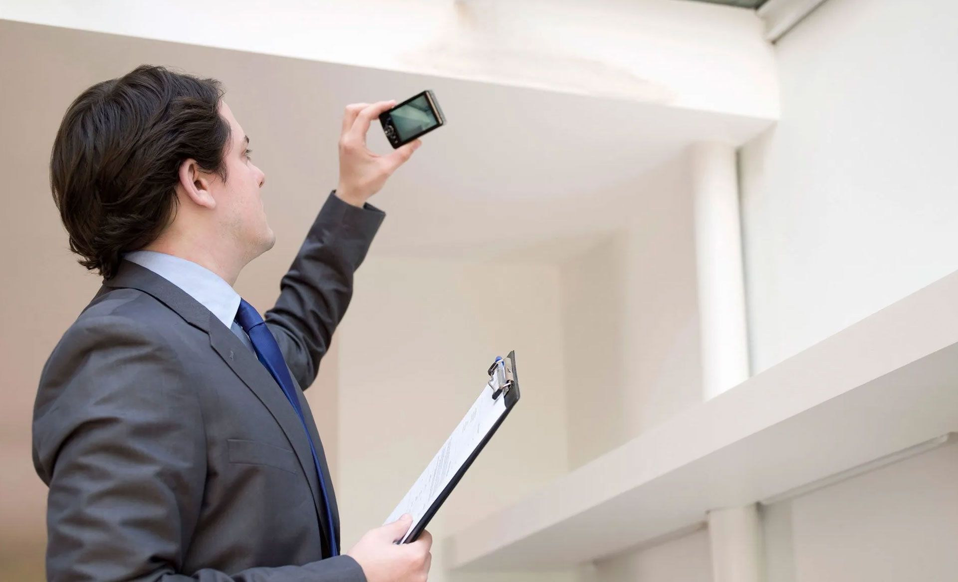 A professional in a suit holds a clipboard and uses a smartphone to take a photo of an indoor ceiling.
