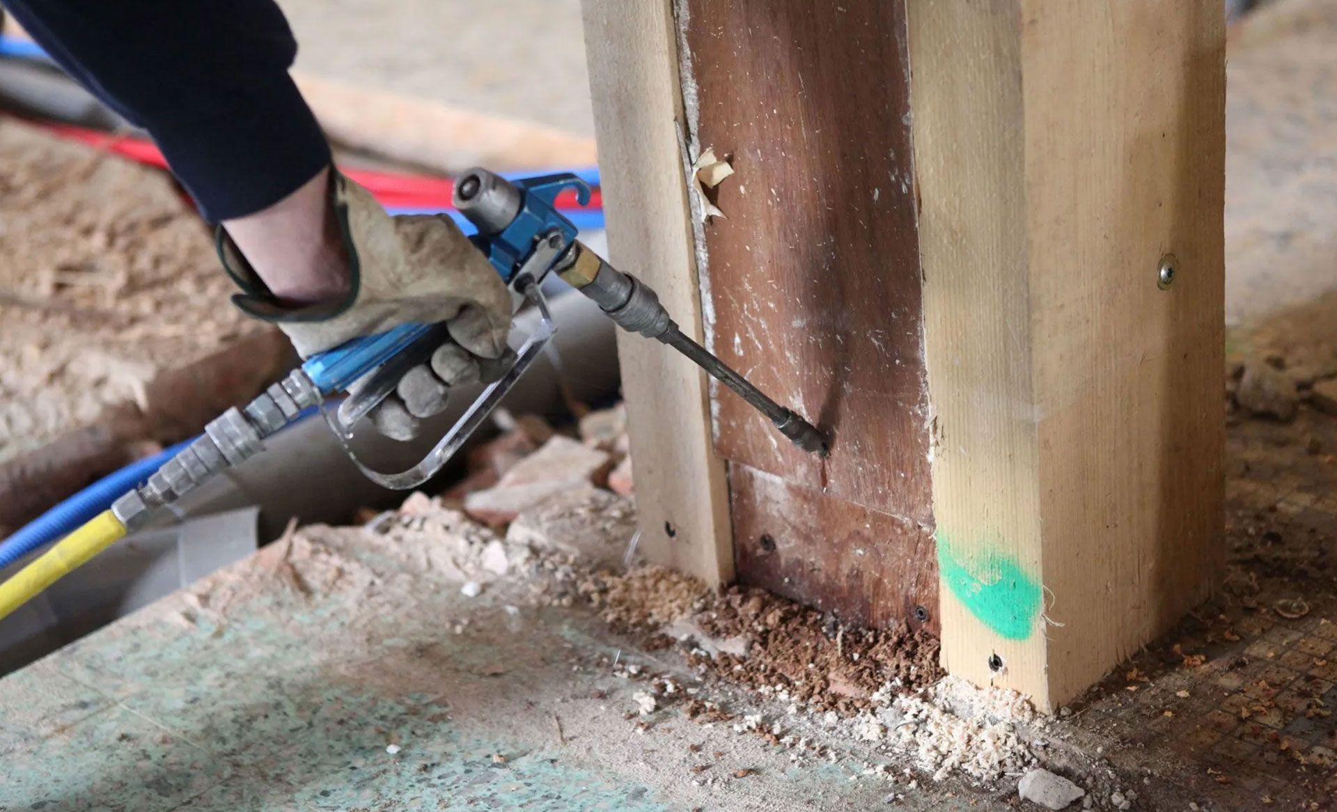 A gloved hand uses a pneumatic spray tool to inject material into a wooden post on a construction site.