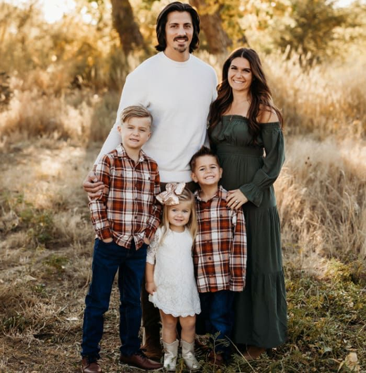 Family of five standing together in a sunlit grassy field, wearing coordinated autumn clothing.