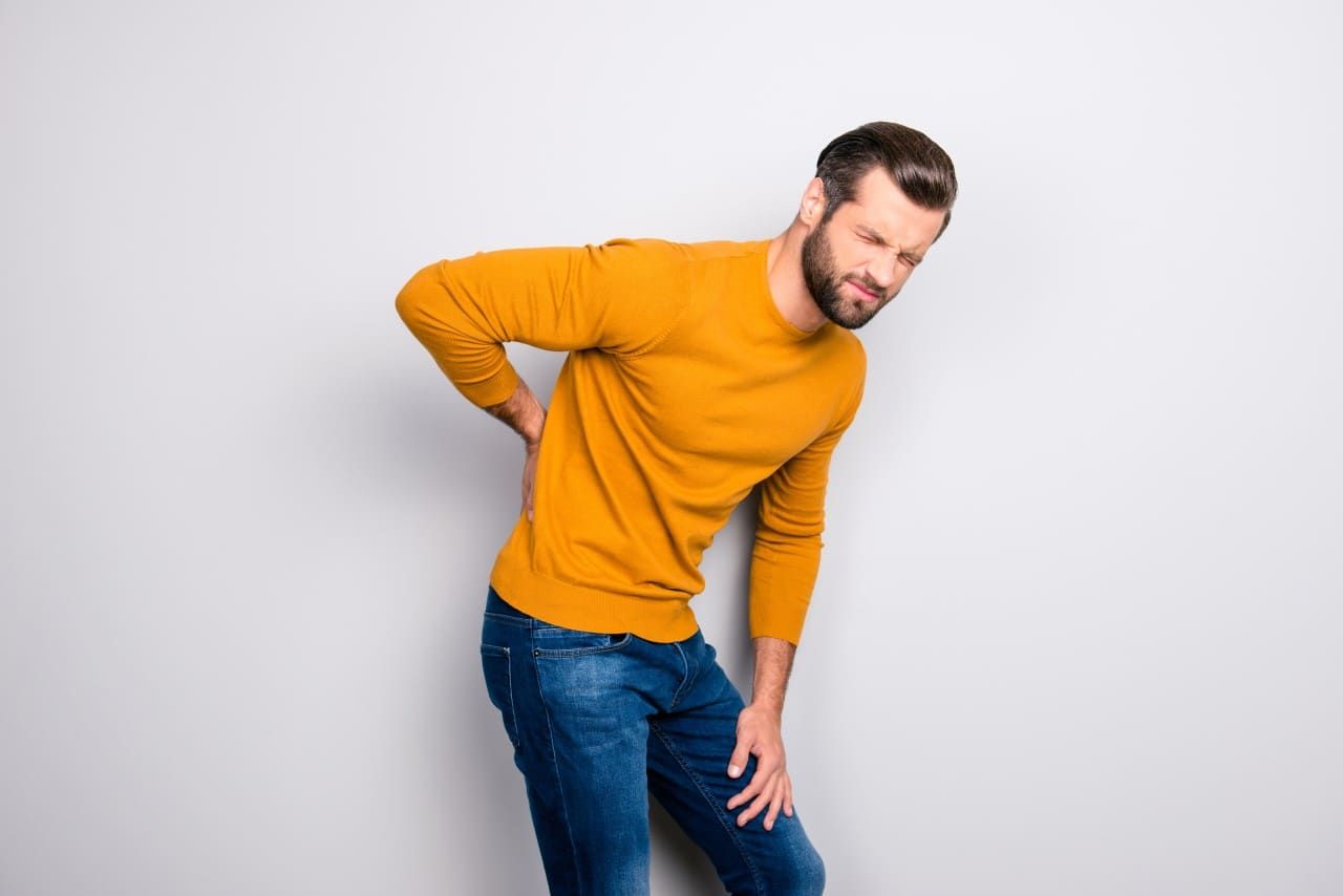 Man in an orange sweater clutching his lower back, bending forward in pain against a plain gray background