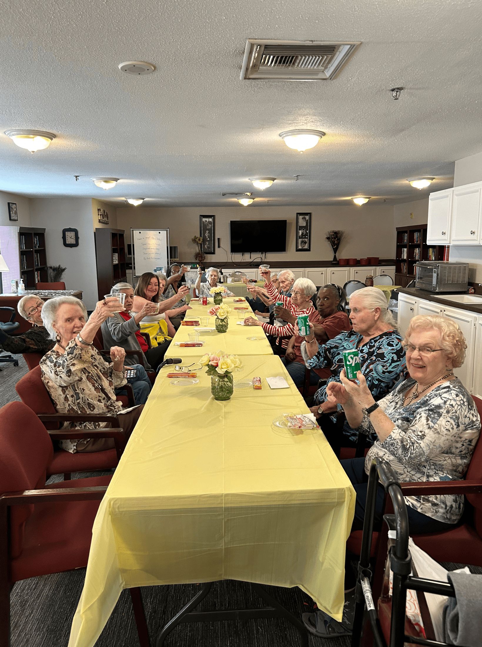 People at a long table, toasting drinks. Yellow tablecloth. Interior shot with a television and decor.