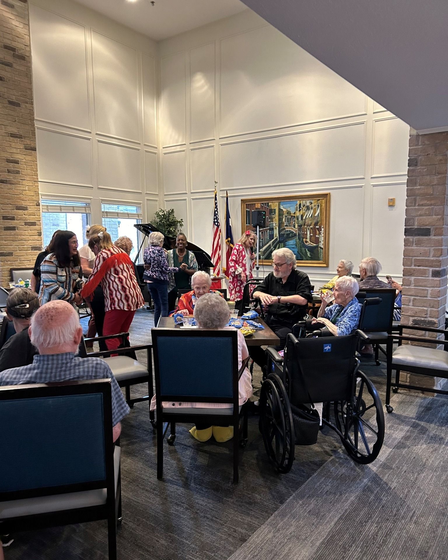 Group of seniors in a room. Some seated, some standing. One person in a red and white patterned jacket. Flags in the background.