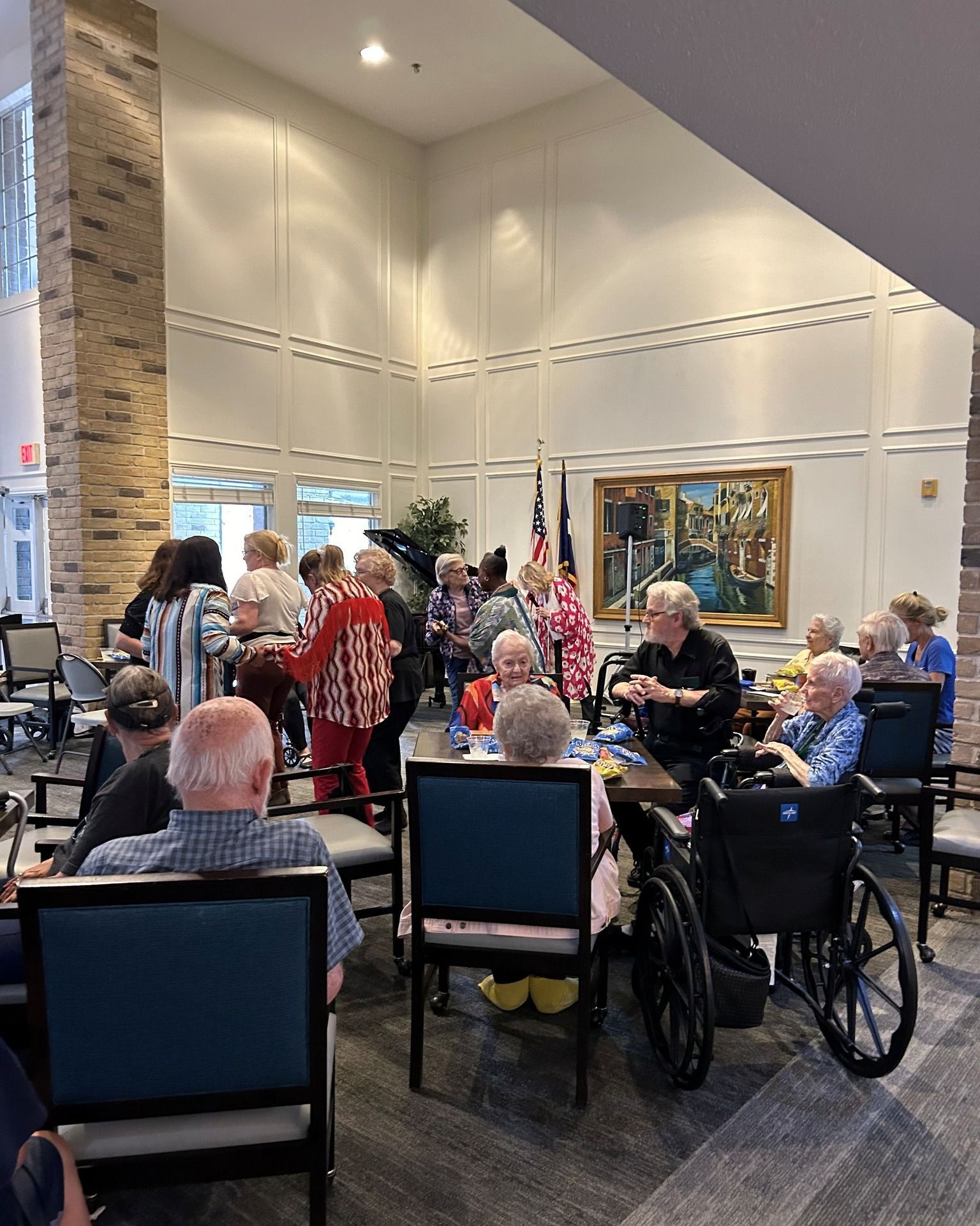 Group of seniors at an event, some dancing. Room with high ceilings, art, and flag. People in chairs, some in wheelchairs.