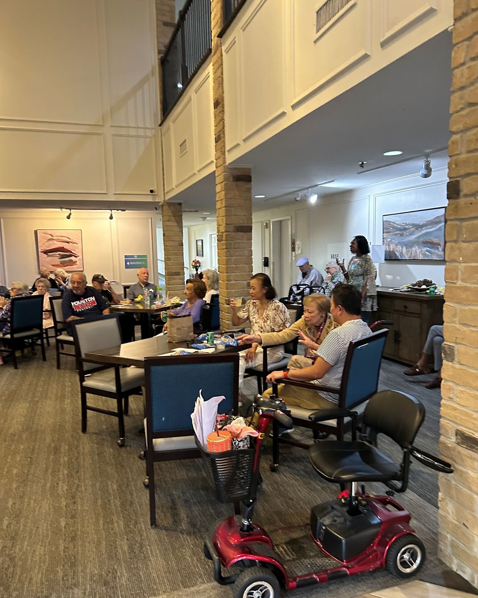 People seated at tables in a room, some interacting. A mobility scooter is in the foreground.