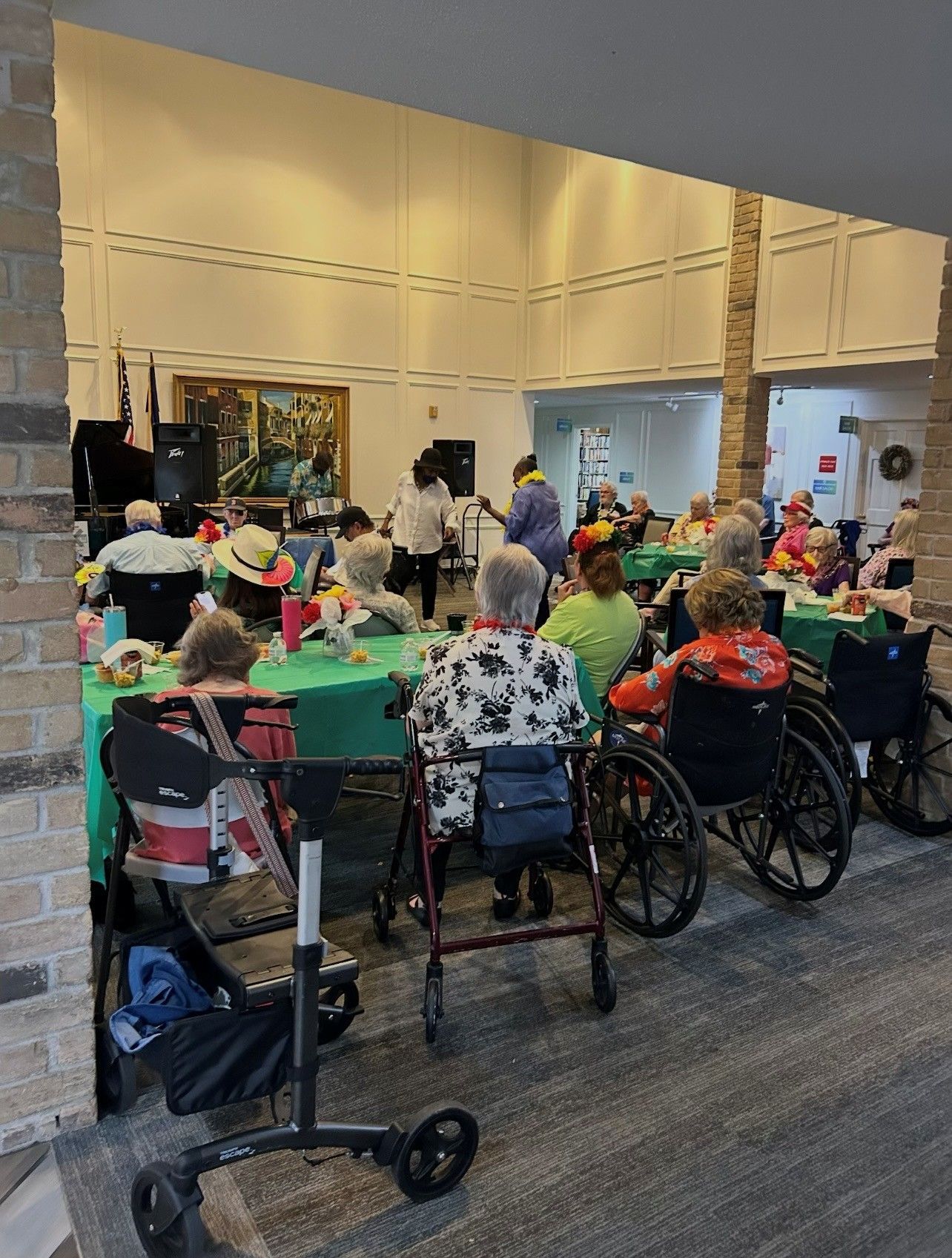 People with walkers and wheelchairs gather at green-covered tables for an indoor event with a speaker.