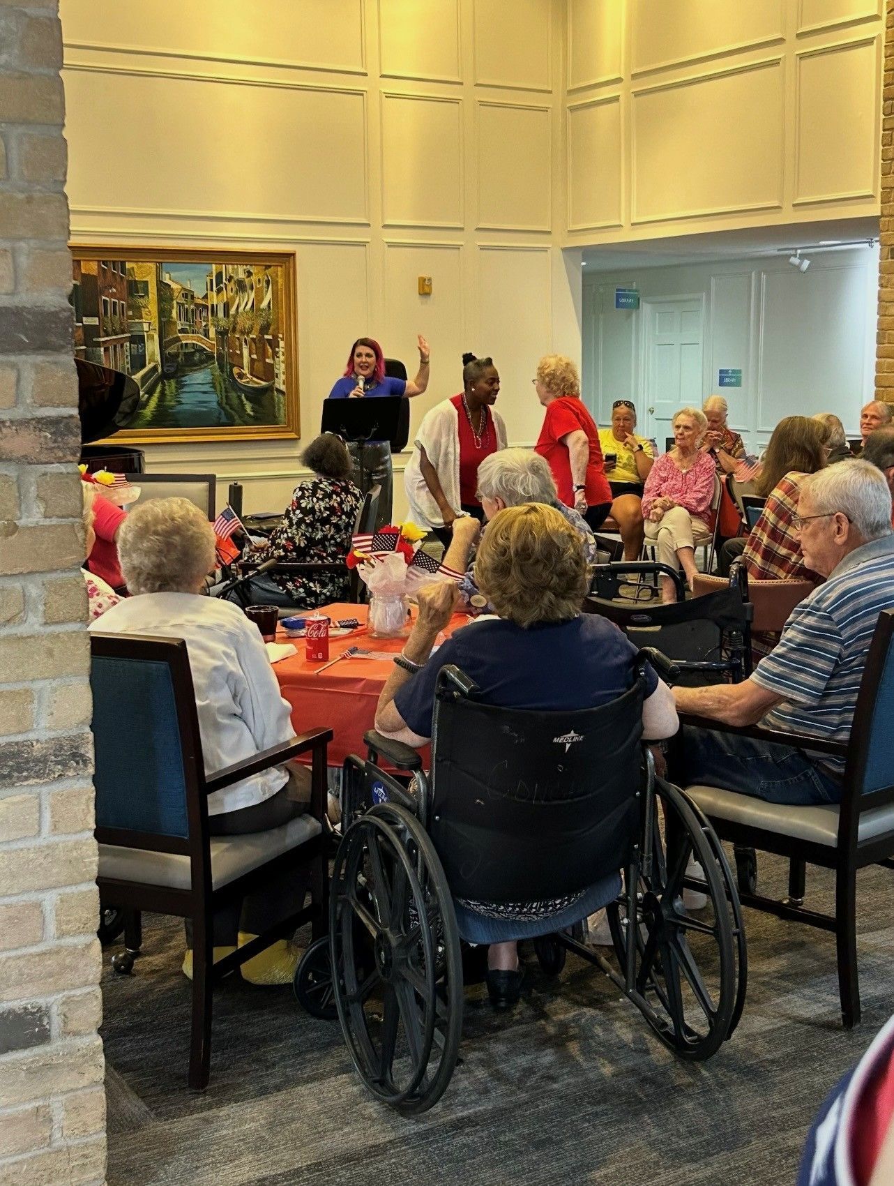 People sit at tables in a room with a large painting while a speaker stands at a podium in the background.