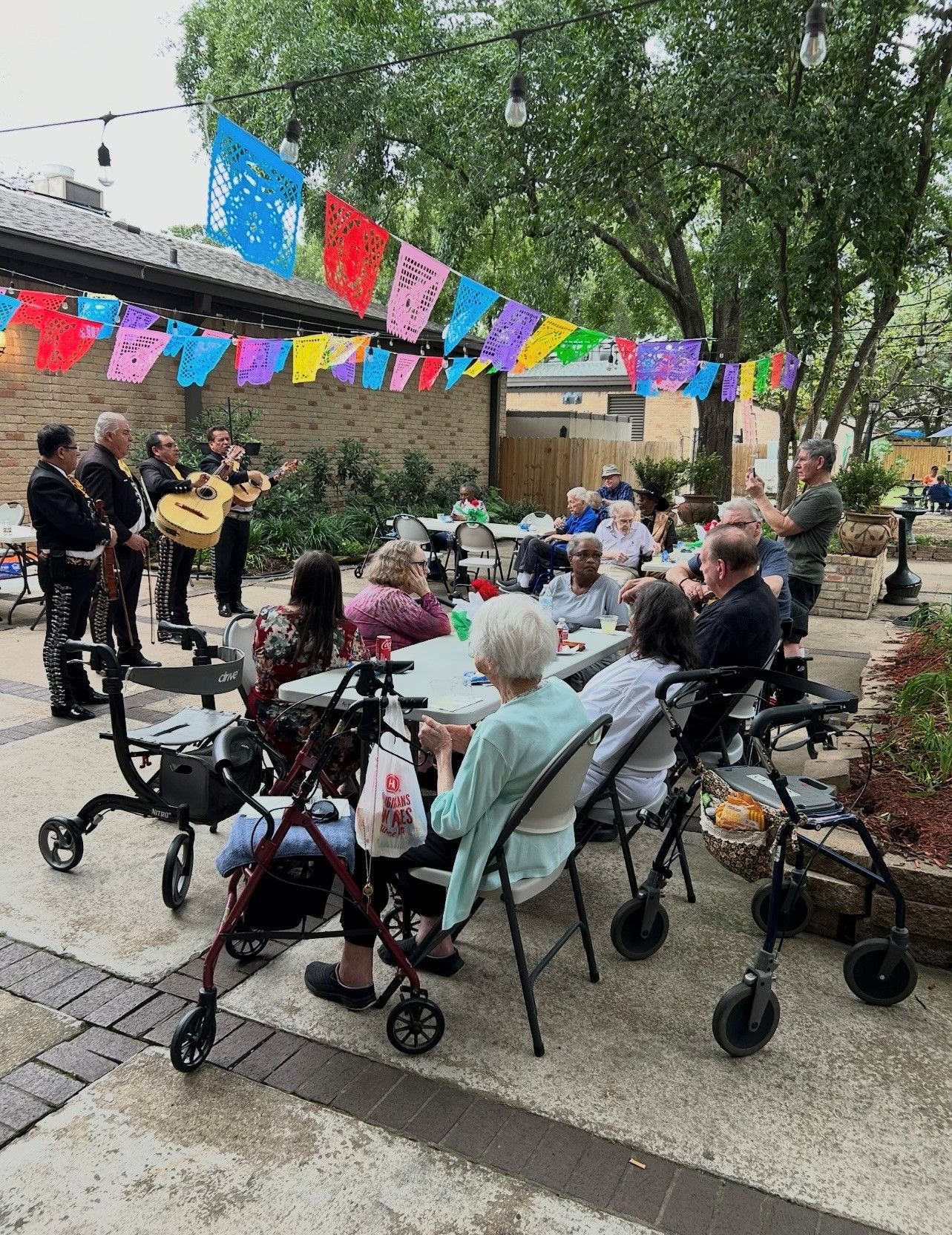 Mariachi band performing outdoors for a group of seated people, under colorful festive banners in a patio setting.
