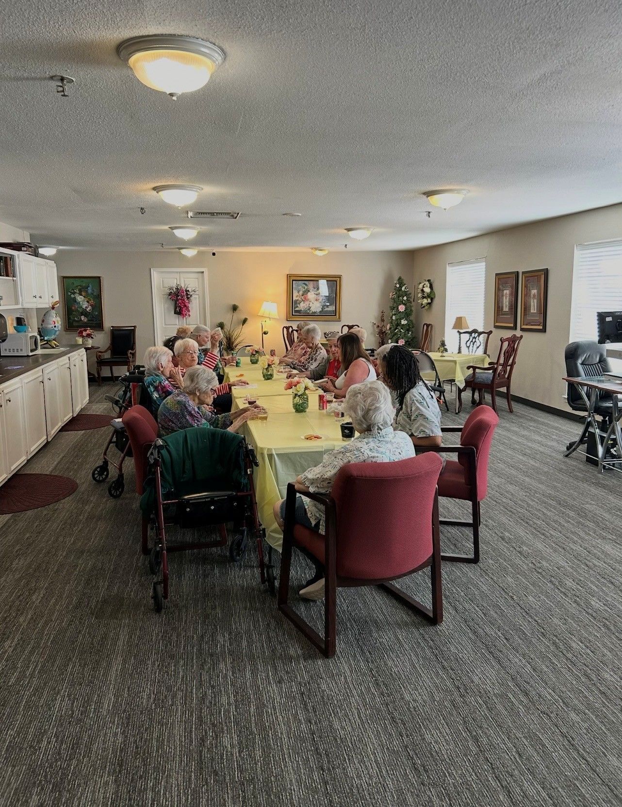 People sit at a long yellow dining table in a community room with gray carpet and overhead lighting.