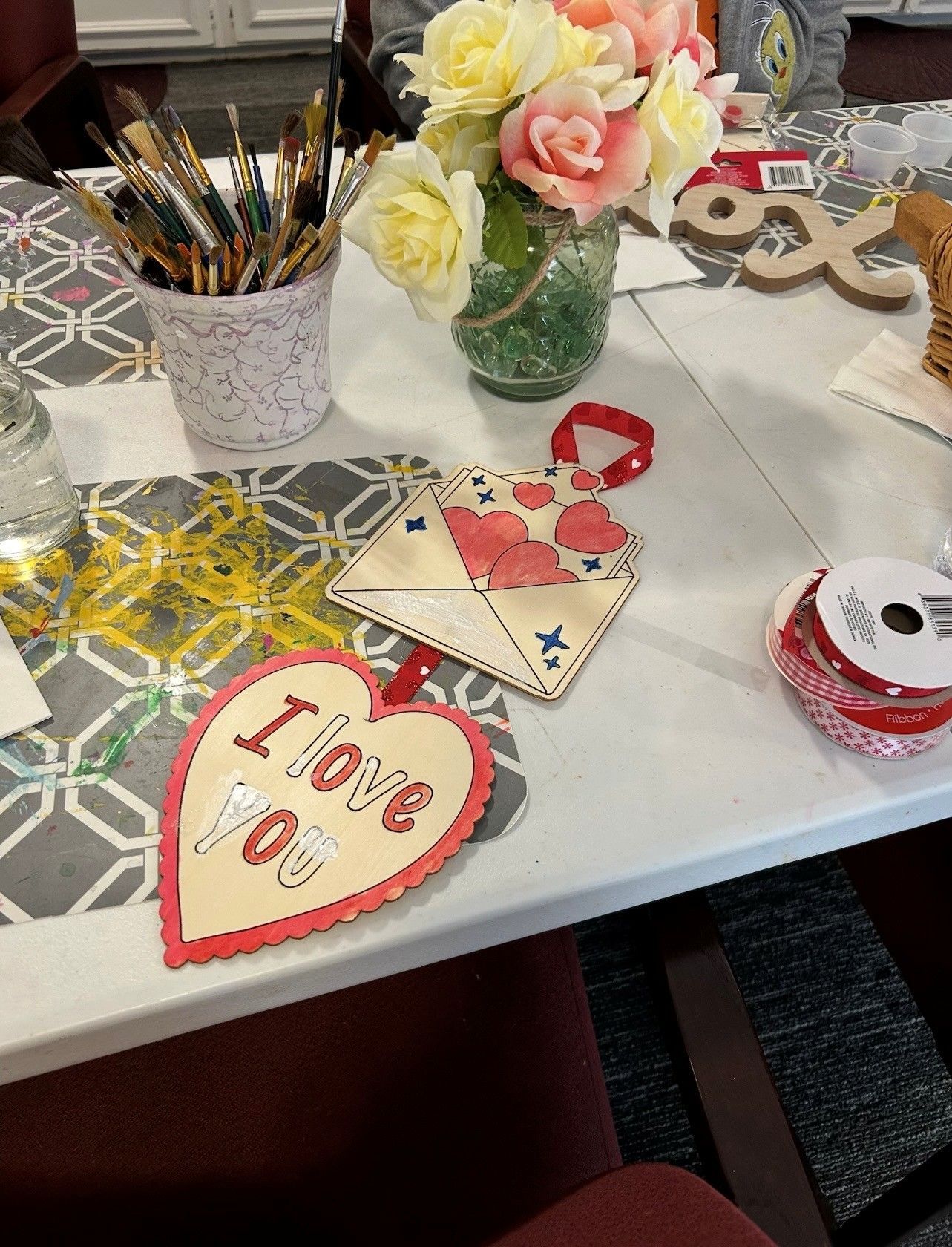 Crafts with hearts and an envelope on a table next to a paint brush holder and a bouquet of flowers.