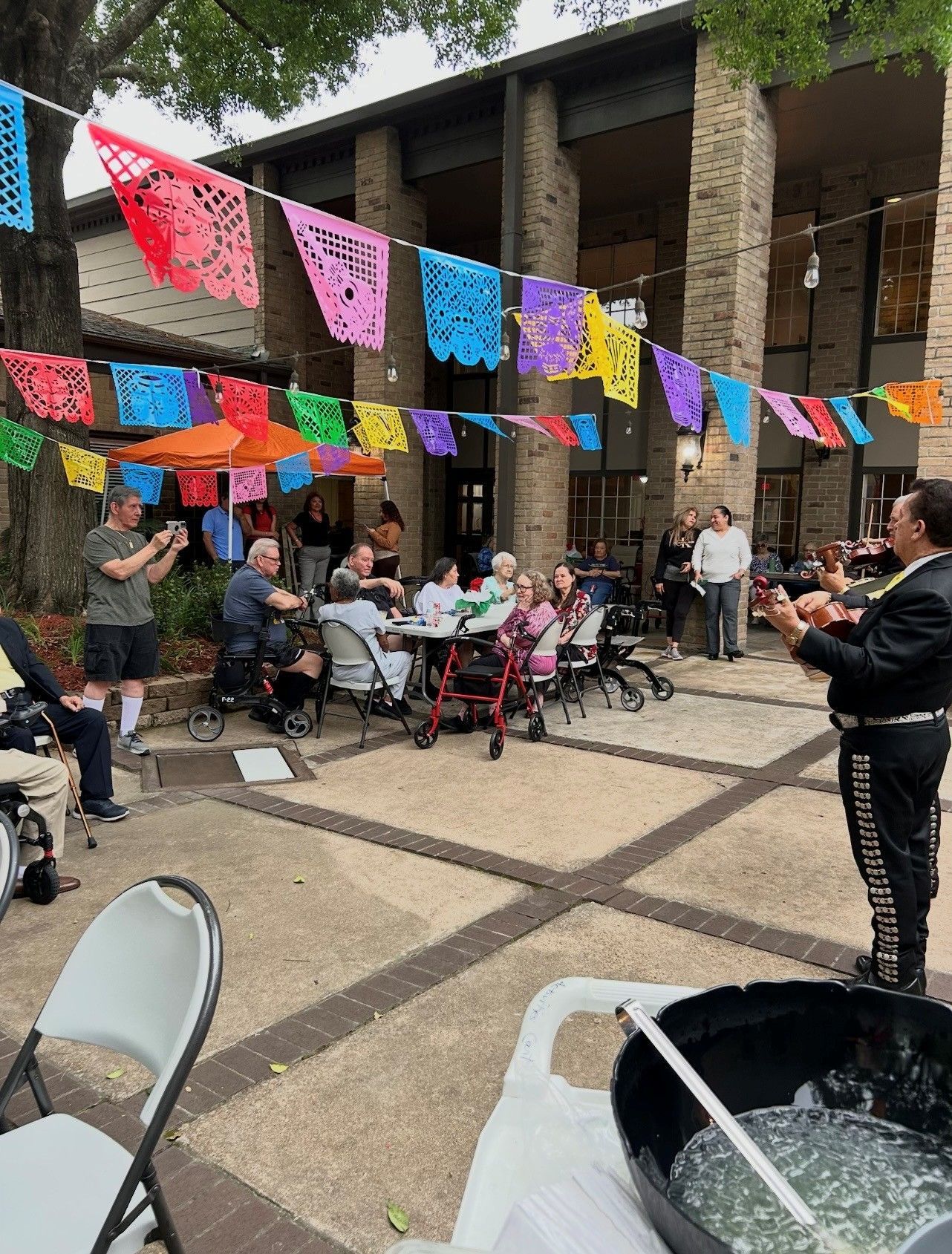 Outdoor gathering with colorful papel picado bunting, seated attendees, and a performer in a black suit.
