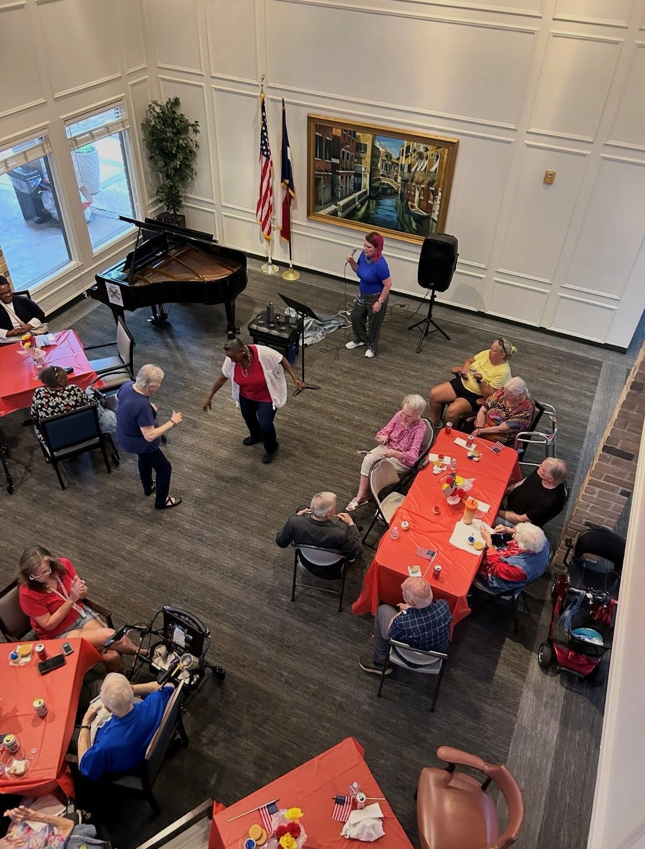 A group of people gathering for a social event in a community hall with tables, a piano, and a person leading an activity.